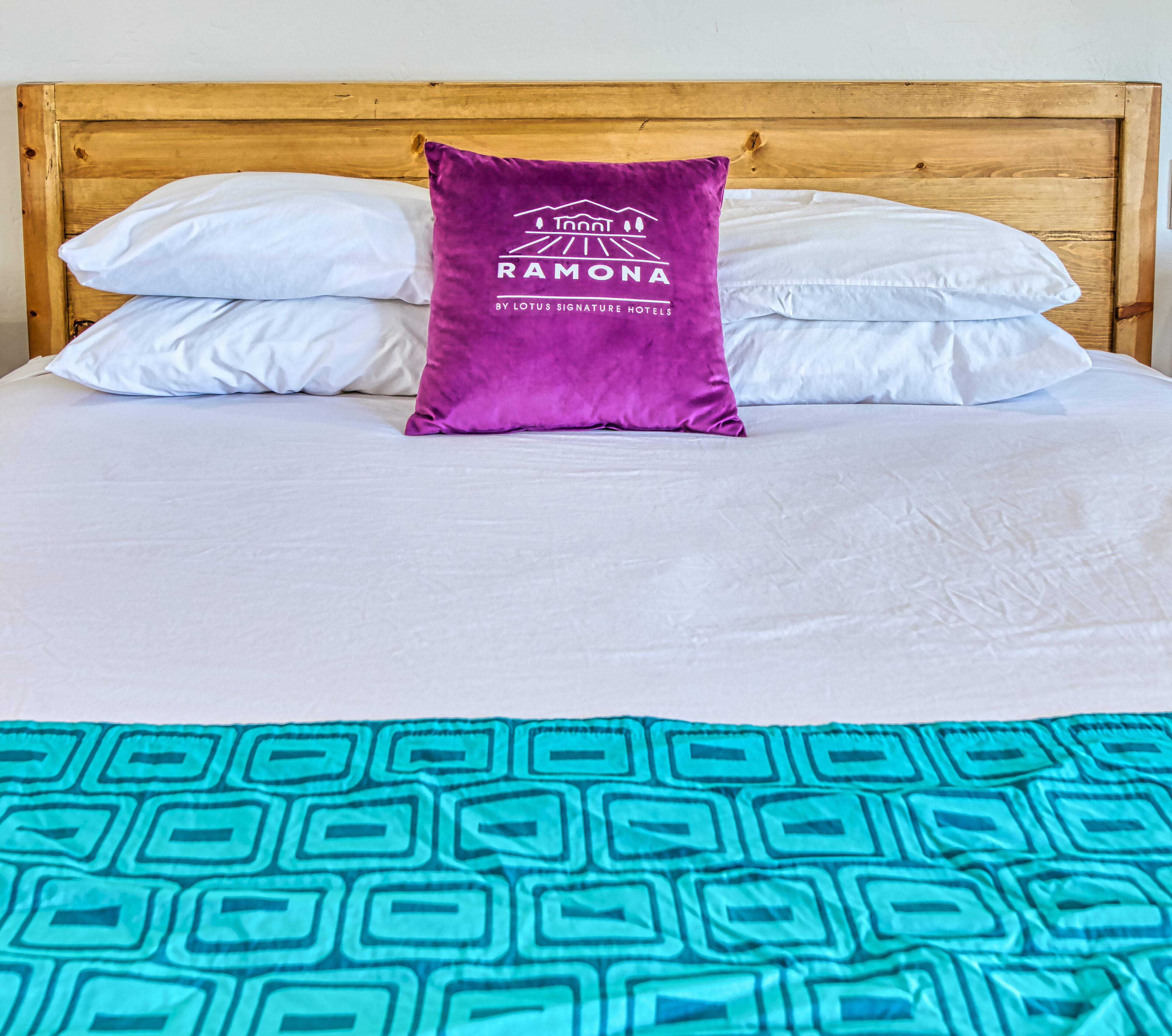 Uma cama com lençóis brancos, um travesseiro roxo com um logotipo e um cobertor estampado em tons de verde-água.