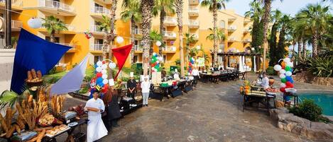 An outdoor dining area with food stalls, palm trees, and a pool.