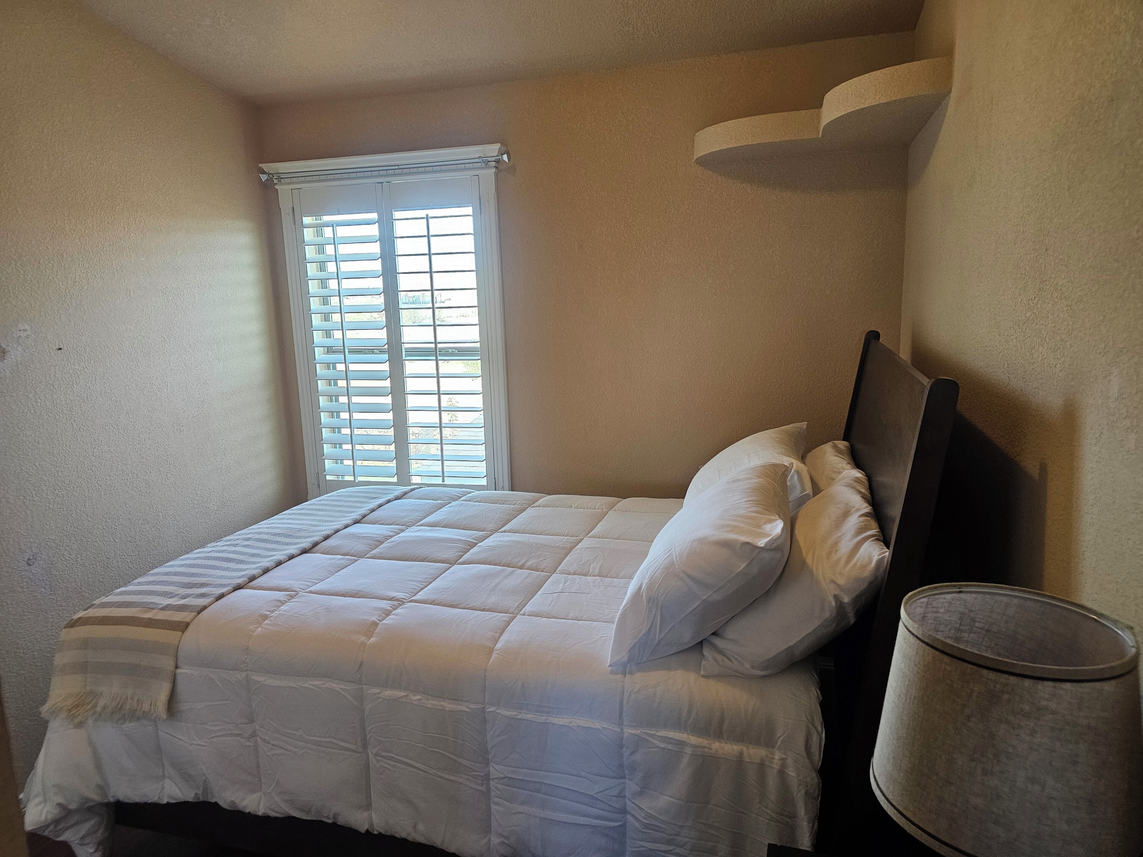 Ein Schlafzimmer mit Bett, Fenster mit Jalousien und einer Lampe.