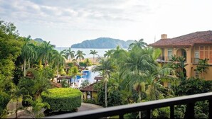 Un complexe hôtelier doté d’une piscine entourée d’une végétation luxuriante et de palmiers, offrant une vue sur une plage et un paysage montagneux en arrière-plan.