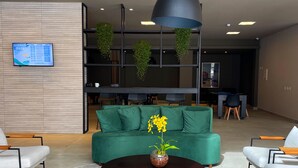 Una moderna zona de recepción con un sofá verde, una mesa de centro con flores, una planta colgante y una pantalla digital en la pared.