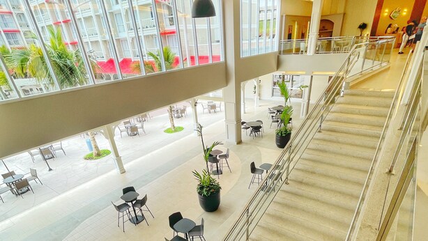 Amplio espacio interior con varios niveles, que incluye una escalera central, mesas de comedor y plantas en macetas.