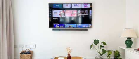 Ein Wohnzimmer mit einem an der Wand montierten Flachbildfernseher, der verschiedene Kanäle zeigt, einem Glastisch mit Büchern und einer Vase, einem Korbwäschesbehältnis, einer Topfpflanze und einer Lampe auf einem weißen Nachttisch.