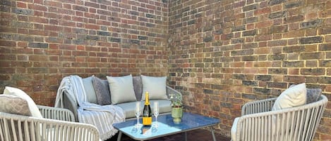 Un'area patio con un muro in mattoni, mobili in vimini, un tavolo in vetro e una bottiglia di champagne.