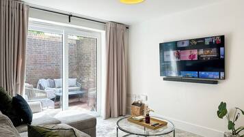 A living room with a sofa, a glass coffee table, a television mounted on the wall, and a sliding glass door leading to an outdoor area.