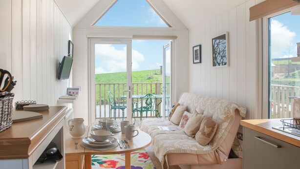 Ein gemütliches Zimmer mit hohen Decken, einem Esstisch mit Stühlen, einer Couch und Blick auf ein grünes Feld durch ein großes Fenster.