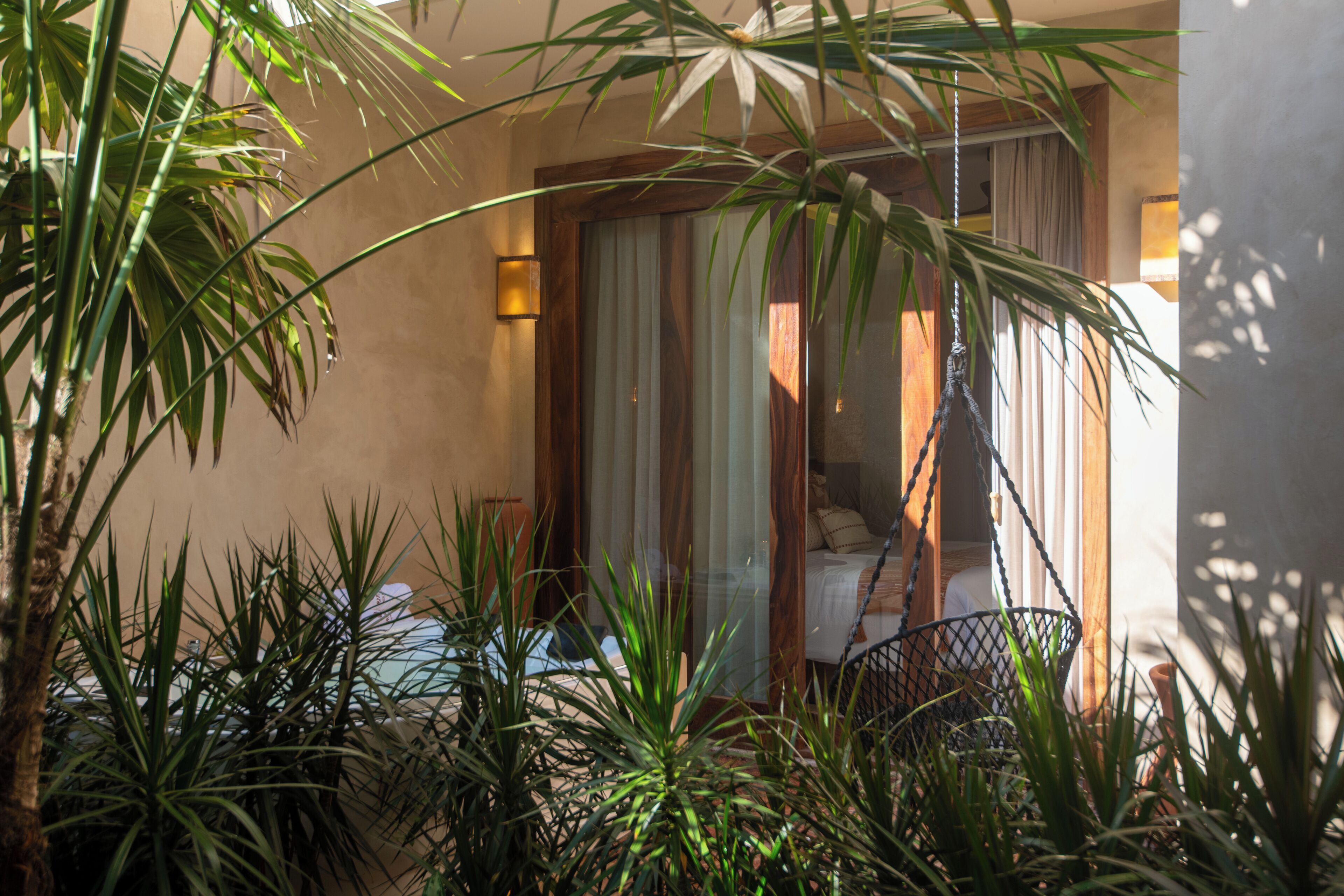 Un balcon avec un fauteuil en osier, entouré de plantes tropicales, et une porte en bois avec des rideaux.