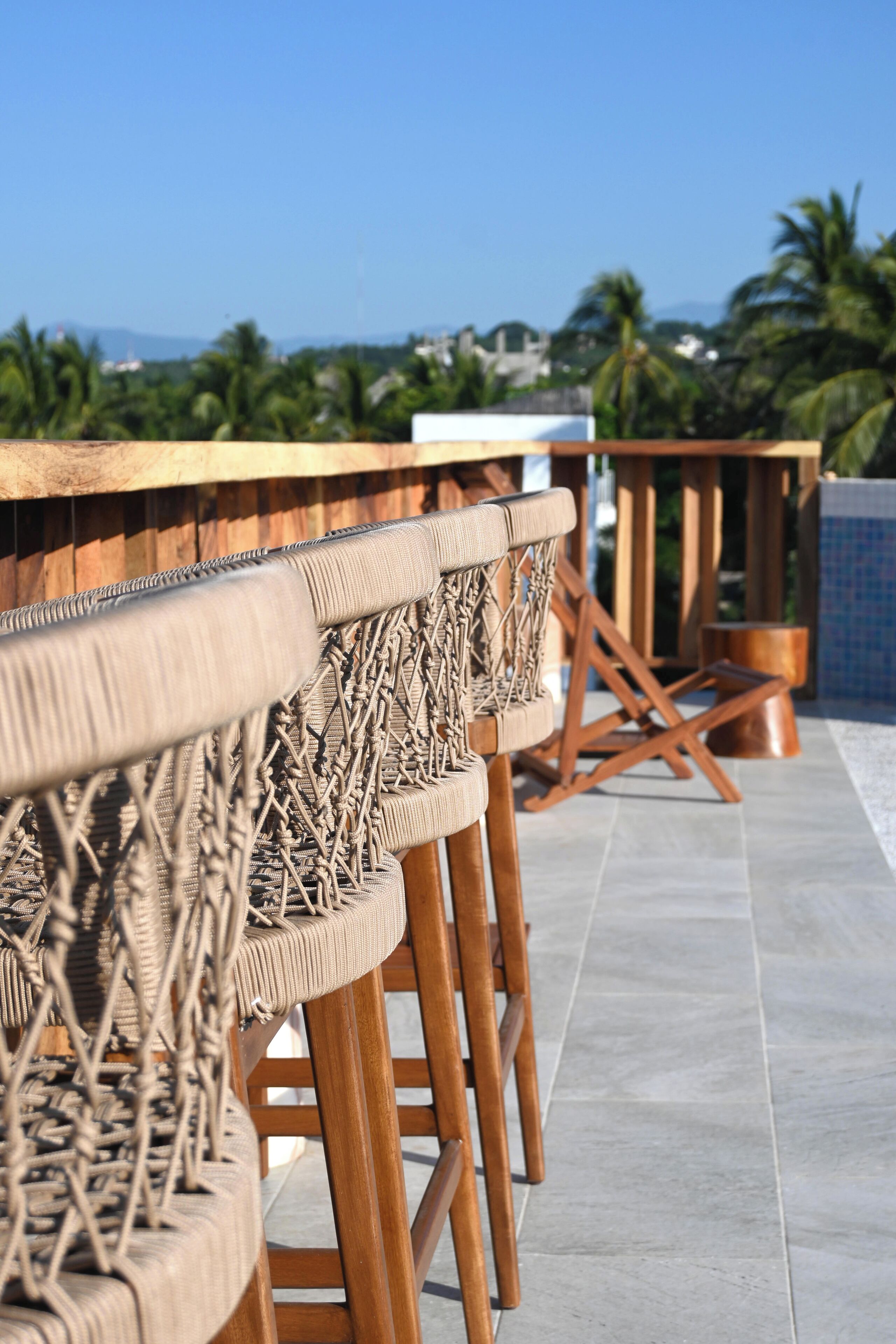 A row of wicker bar stools with wooden legs and a cushioned seat, arranged on a tiled patio with a view of palm trees and a distant mountain range.