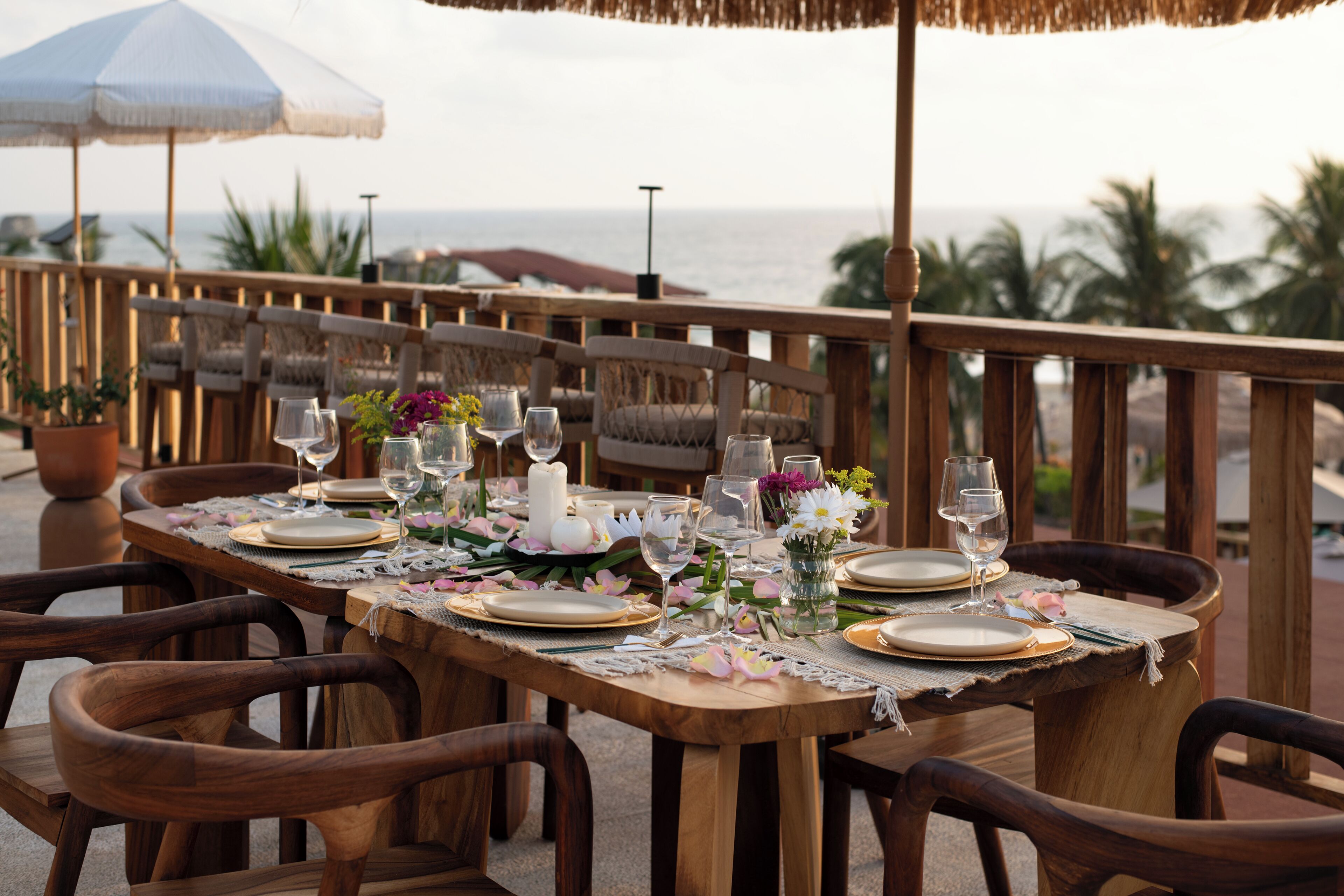 A wooden dining table set with plates, glasses, and flowers, overlooking the ocean.