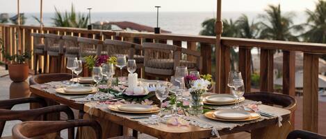 A wooden dining table set with plates, glasses, and flowers, overlooking the ocean.