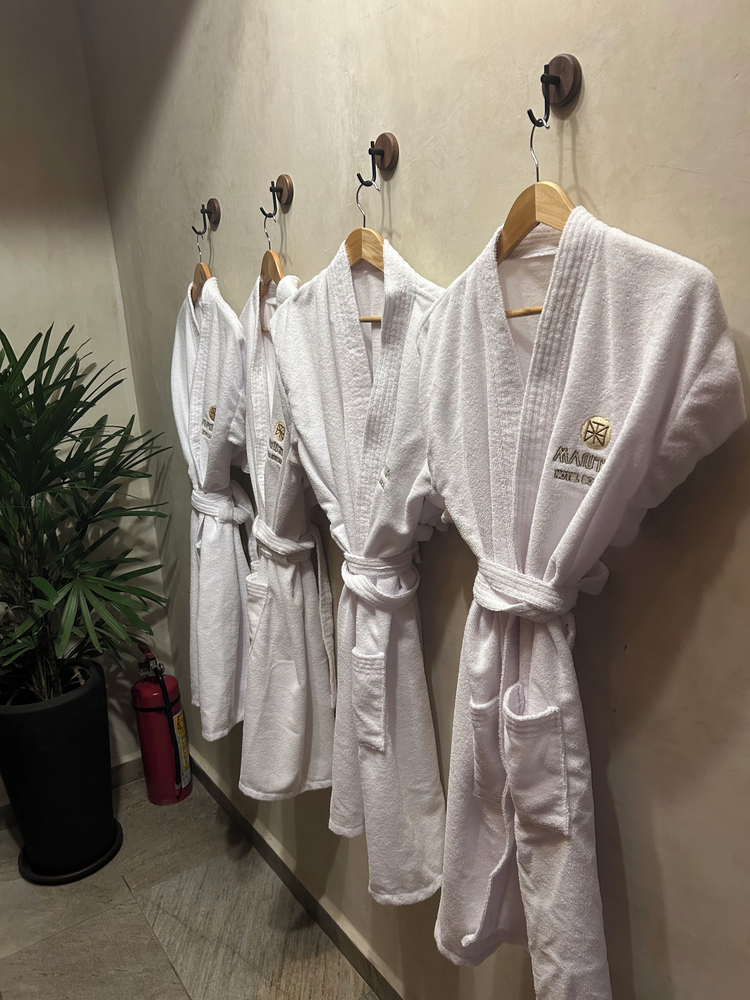 A row of white robes hanging on hooks against a wall, with a potted plant and a fire extinguisher nearby.