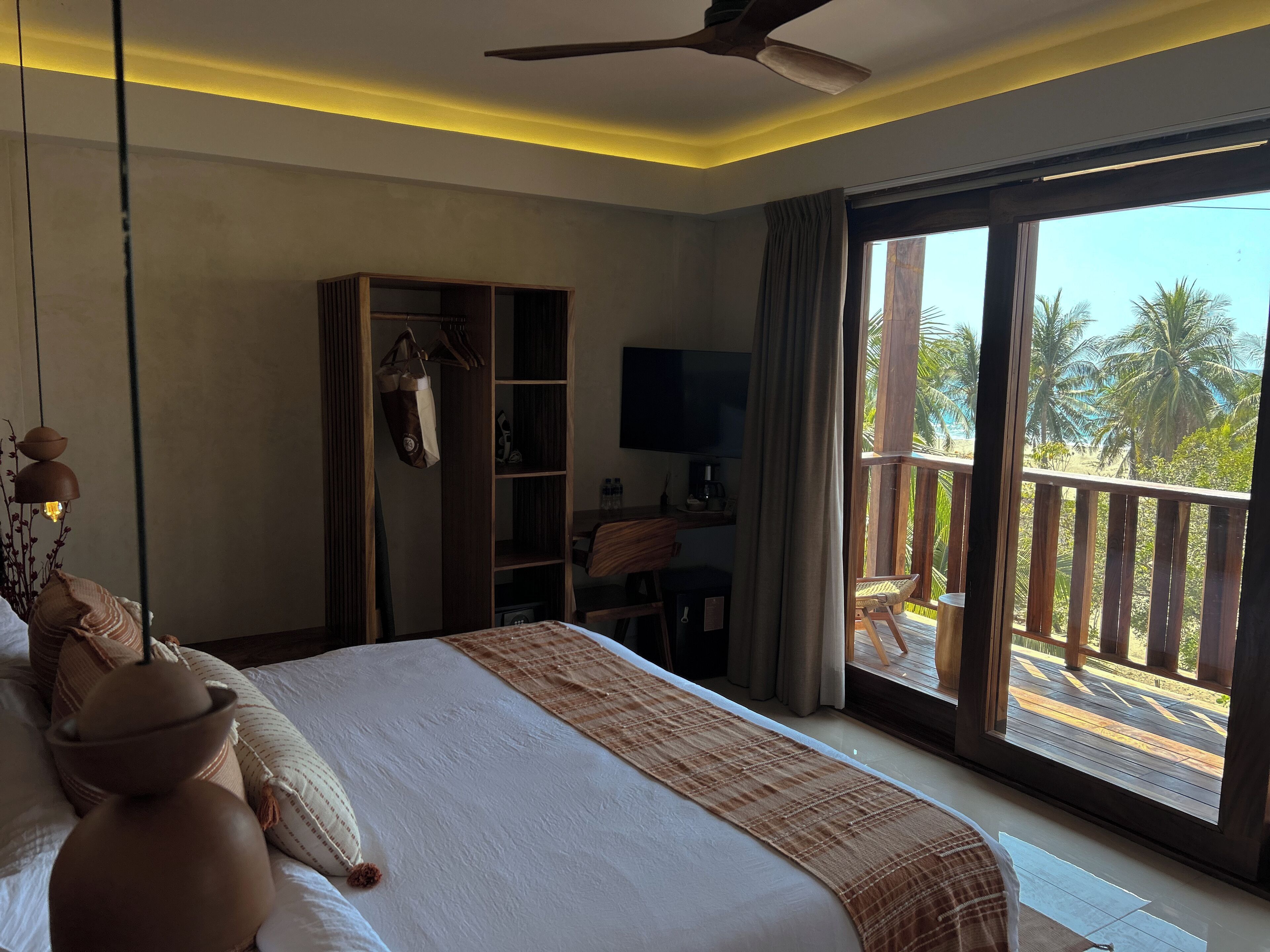 Habitación de hotel con cama, armario, escritorio con silla, televisor y balcón con vista a palmeras y la playa.