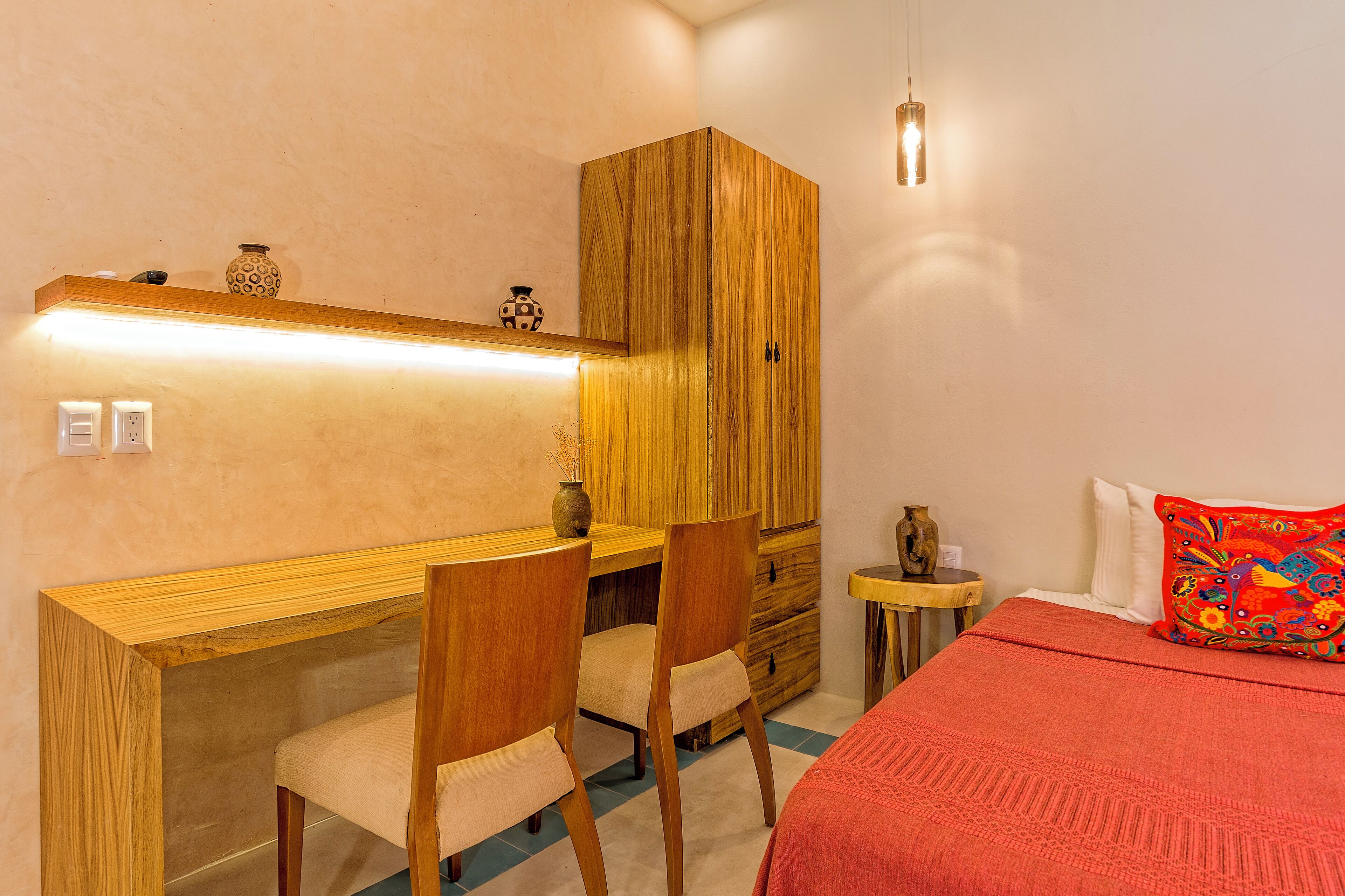 A bedroom with a wooden desk, two chairs, a bed with a red blanket, and a small bedside table with a vase.
