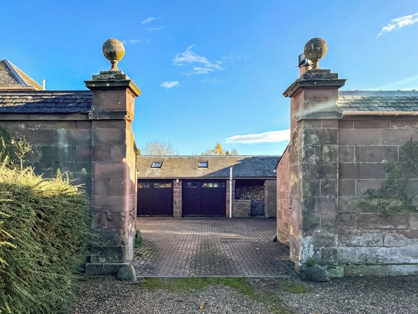 Une entrée fermée par un portail, avec des piliers en pierre et un chemin pavé menant à un bâtiment doté de deux portes de couleur sombre.
