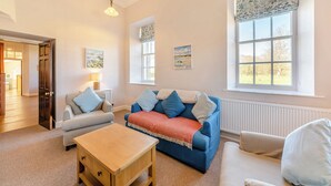 A living room with a blue sofa, a wooden coffee table, a white armchair, and a door leading to another room. - The Old Bakery - UK48319 (Berwick-Upon-Tweed)
