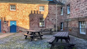 A stone building with a blue door and windows, outdoor wooden picnic tables, and a cobblestone ground. - The Old Bakery - UK48319 (Berwick-Upon-Tweed)