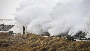 Costa escarpada con olas rompiendo sobre las rocas, una persona de pie en la orilla y un banco en primer plano.