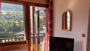 Une pièce comprenant une commode en bois, un téléviseur, un lit confortable recouvert d’une couverture à motifs rouges et blancs, une fenêtre donnant sur un paysage verdoyant et une porte-fenêtre en bois donnant sur un balcon.