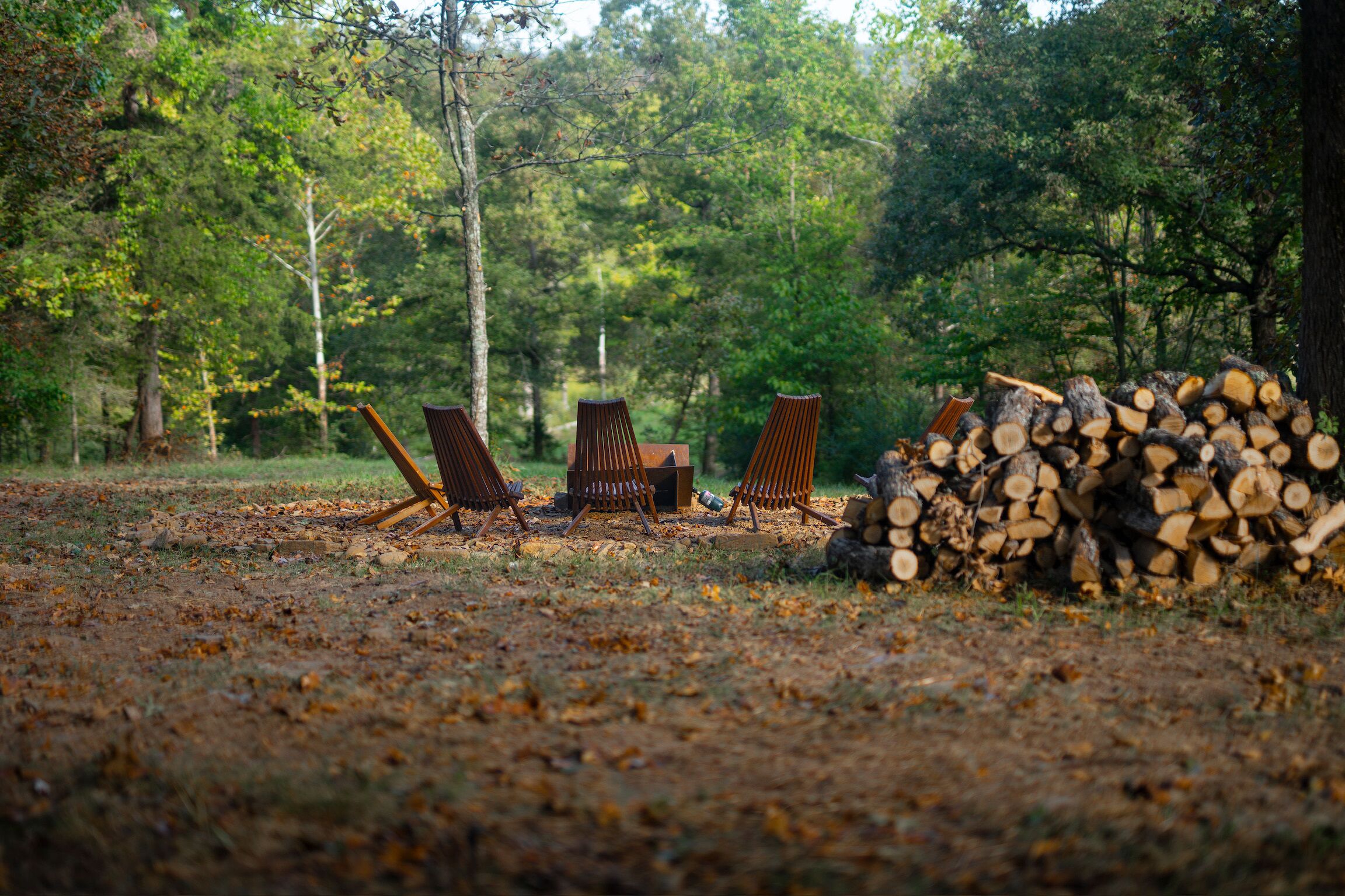 A forest clearing with three wooden chairs and a stack of firewood.
