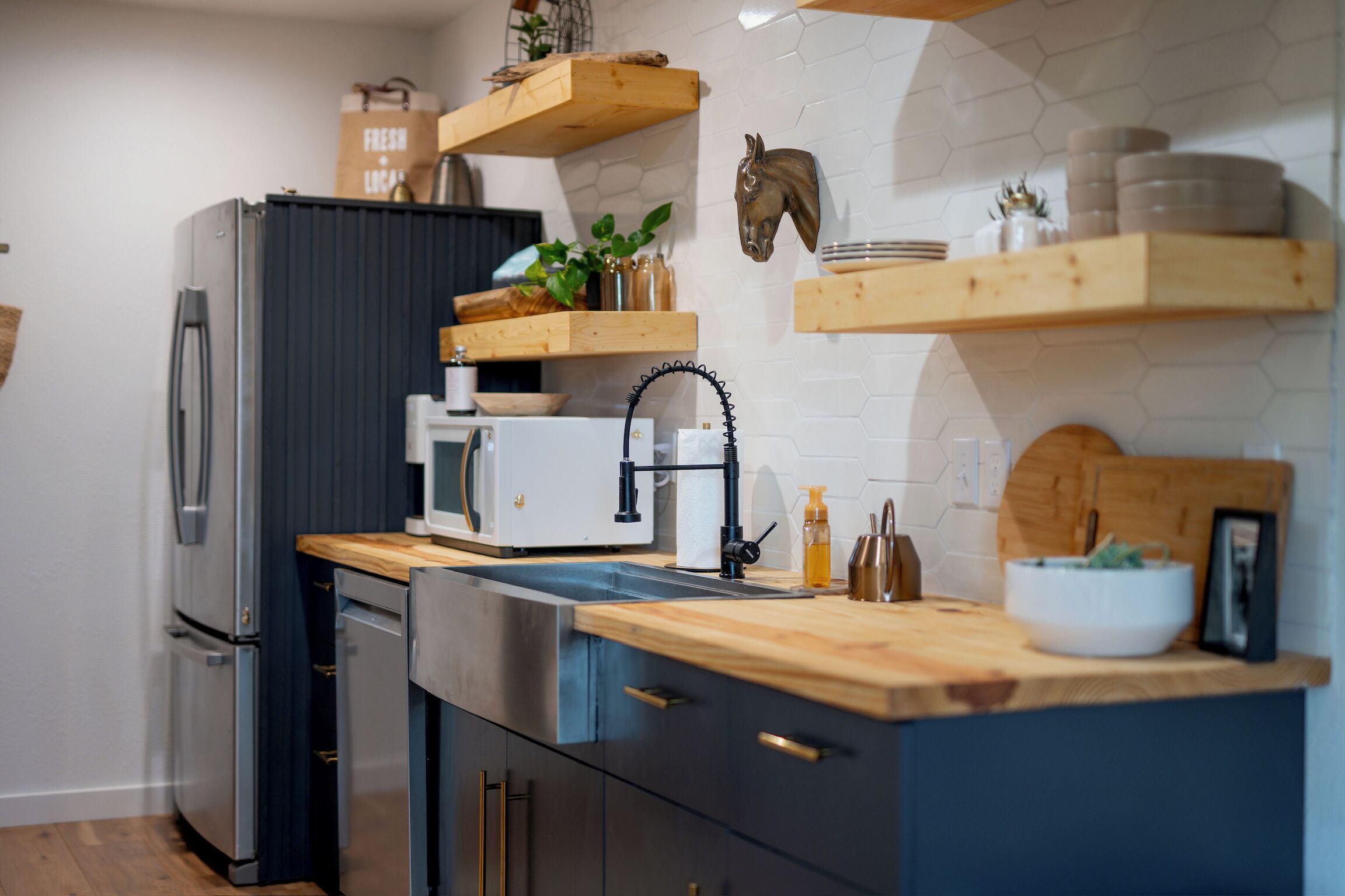 A modern kitchen with a stainless steel refrigerator, a microwave, wooden shelves, and a hexagonal tile backsplash.