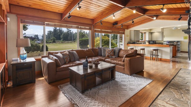 A spacious living area with a wooden ceiling, large windows offering a view of a green landscape, a brown leather sofa, a coffee table, and a kitchen in the background. - Rainier Ranch & Poolside Escape (Enumclaw)