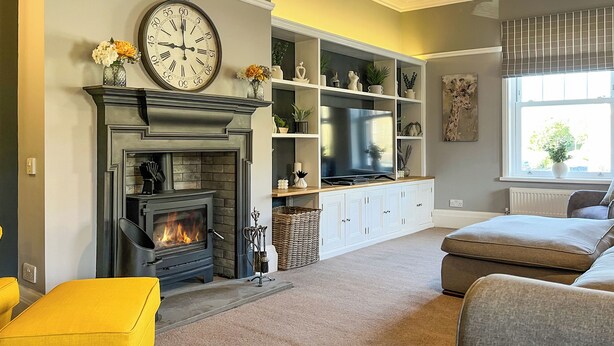 A living room with a fireplace, a large clock, a television, and a window with blinds.