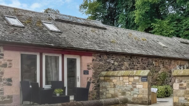 Un bâtiment en pierre avec une façade rose, un petit porche avec des chaises et une entrée surmontée d’un mur en pierre.