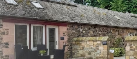 Un edificio de piedra con fachada rosa, un pequeño porche con sillas y una entrada a través de un muro de piedra.