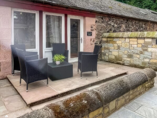 A stone building with a red door and a sign above it. There are two black chairs and a small table with a plant on the patio.