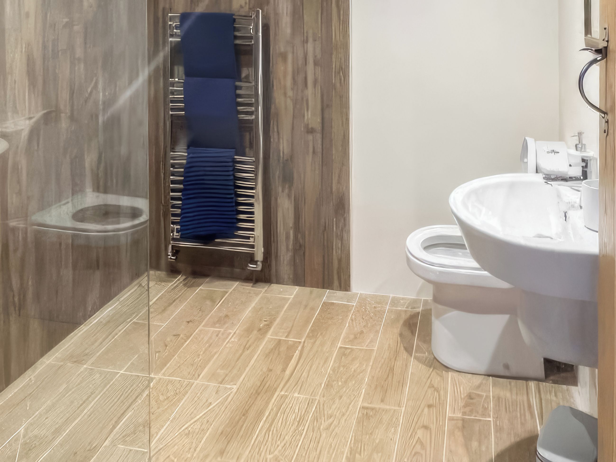 A bathroom with a wooden floor, a glass shower enclosure, a white sink, and a towel rack with a blue towel.
