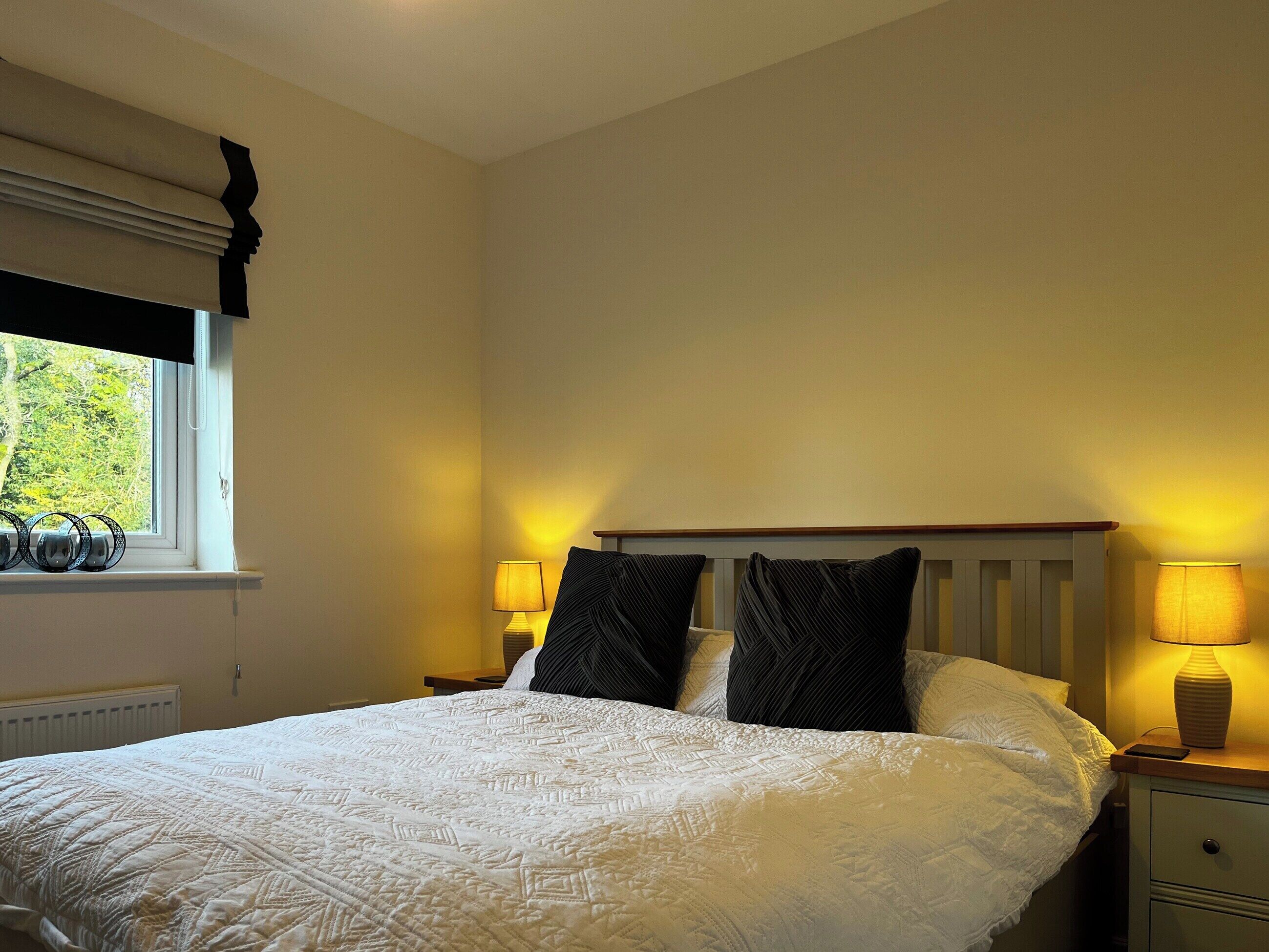 Una cama bien hecha con colcha blanca, dos almohadas negras y cabecera de madera. Hay dos lámparas de noche a cada lado de la cama.