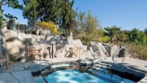 A swimming pool with a stone wall backdrop, two wooden chairs, and a metal ladder.
