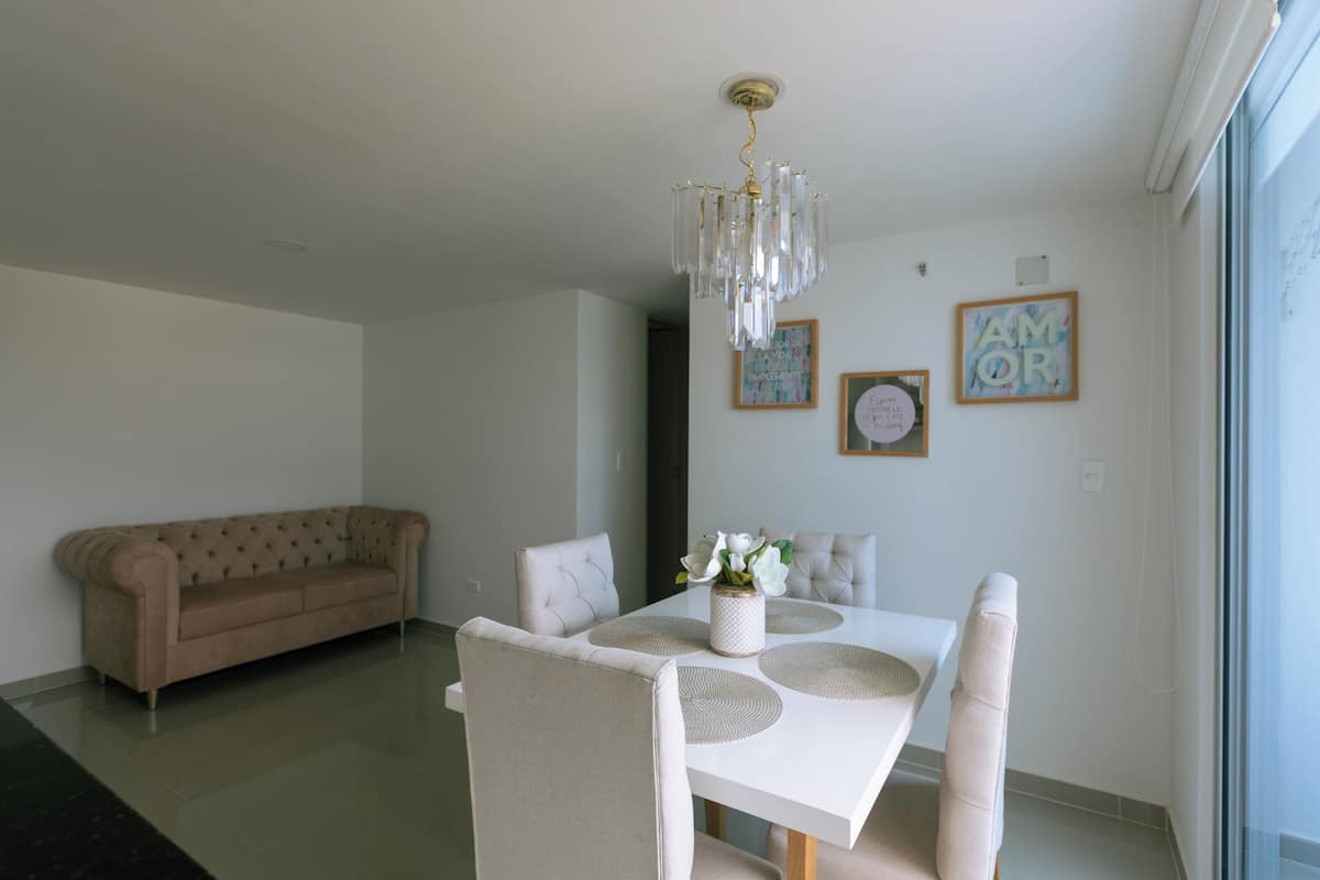 A dining area with a white table, beige chairs, and a chandelier. There is a beige sofa in the background and two framed pictures on the wall.