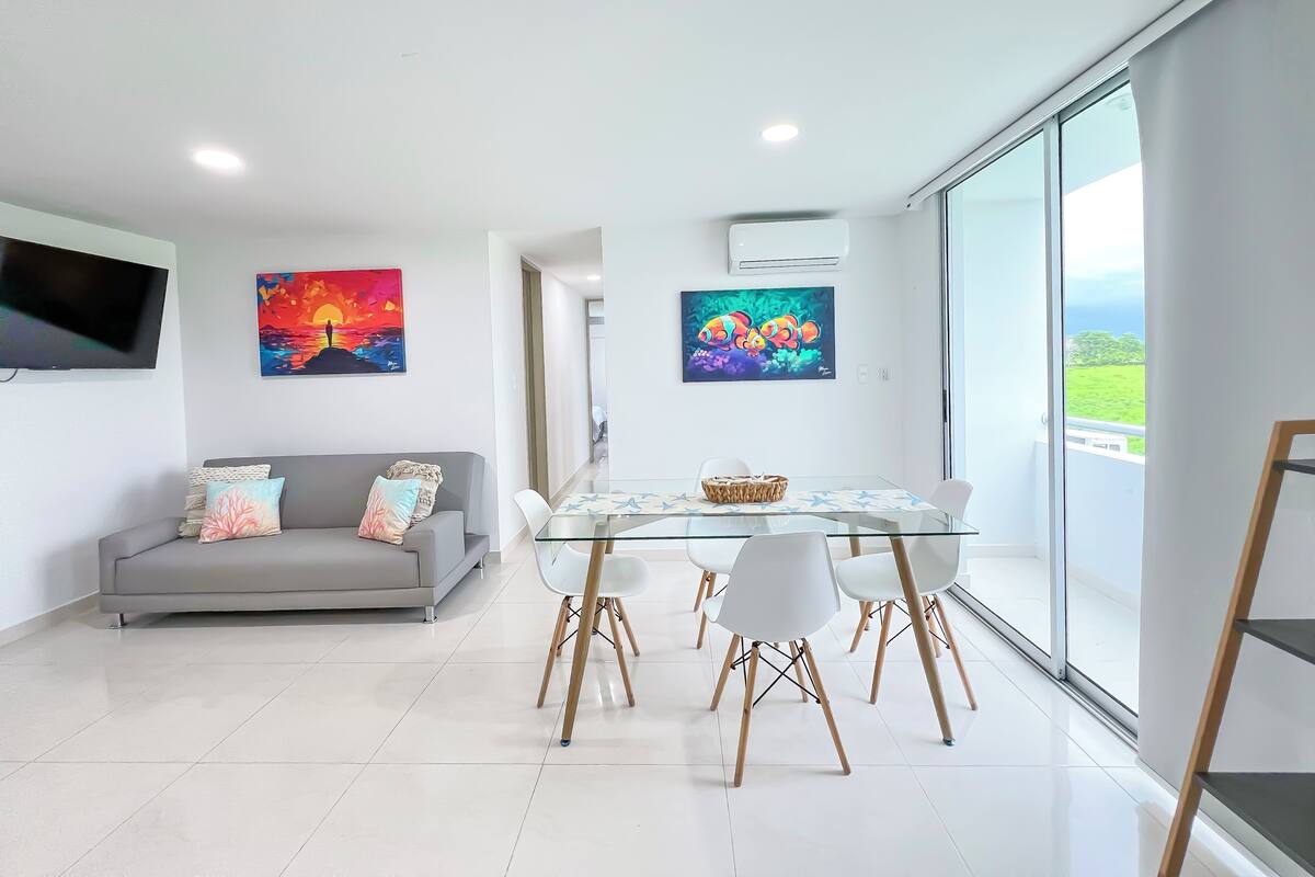 A modern living room with a grey sofa, a glass dining table with chairs, and artwork on the walls.