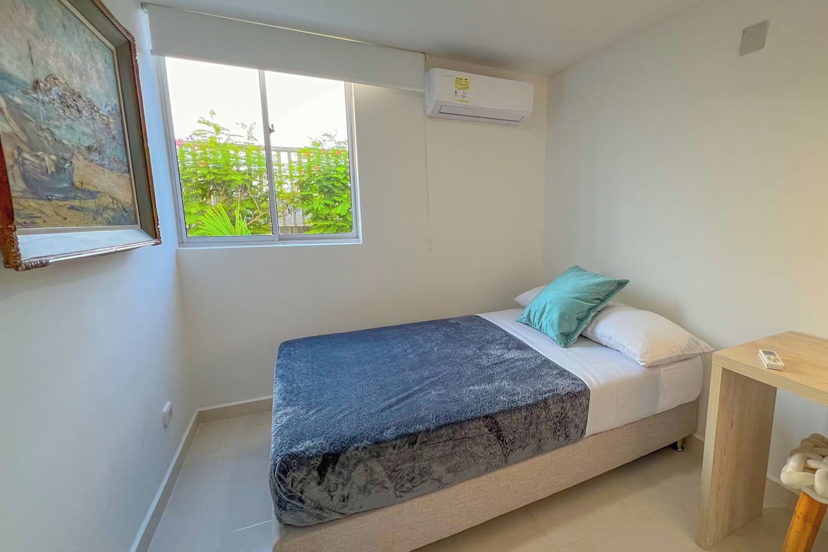 A small, compact room with a bed, a window, an air conditioning unit, a small table, and a chair.