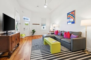 Ein modernes Wohnzimmer mit einem Flachbildfernseher, einer Couch mit bunten Kissen, einem grünen Couchtisch und einem Streifen Teppich.