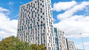 Un moderno grattacielo con una facciata a griglia di finestre, circondato da alberi e altri edifici.