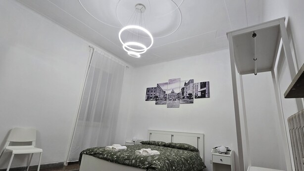 A bedroom with a bed, a chair, a nightstand, a wall-mounted lamp, and a ceiling light fixture. - Optima mansio (Roma)