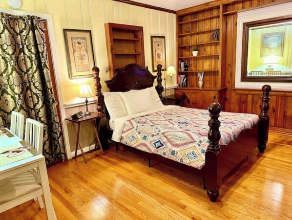 A traditional bedroom with a wooden four-poster bed, a bedside table with a lamp, a small desk, and a bookshelf.