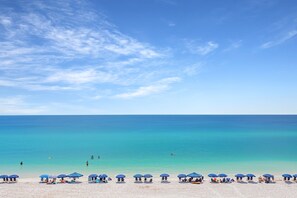 Ein Strand mit klarem türkisblauem Wasser, einem Sandstrand und mehreren blauen Sonnenschirmen.