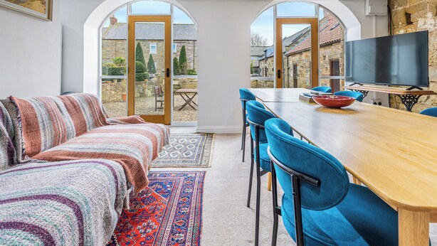 Ein gemütliches Zimmer mit einer Couch, einem Esstisch mit Stühlen, einem Fernseher und großen Fenstern mit Blick auf einen Innenhof.