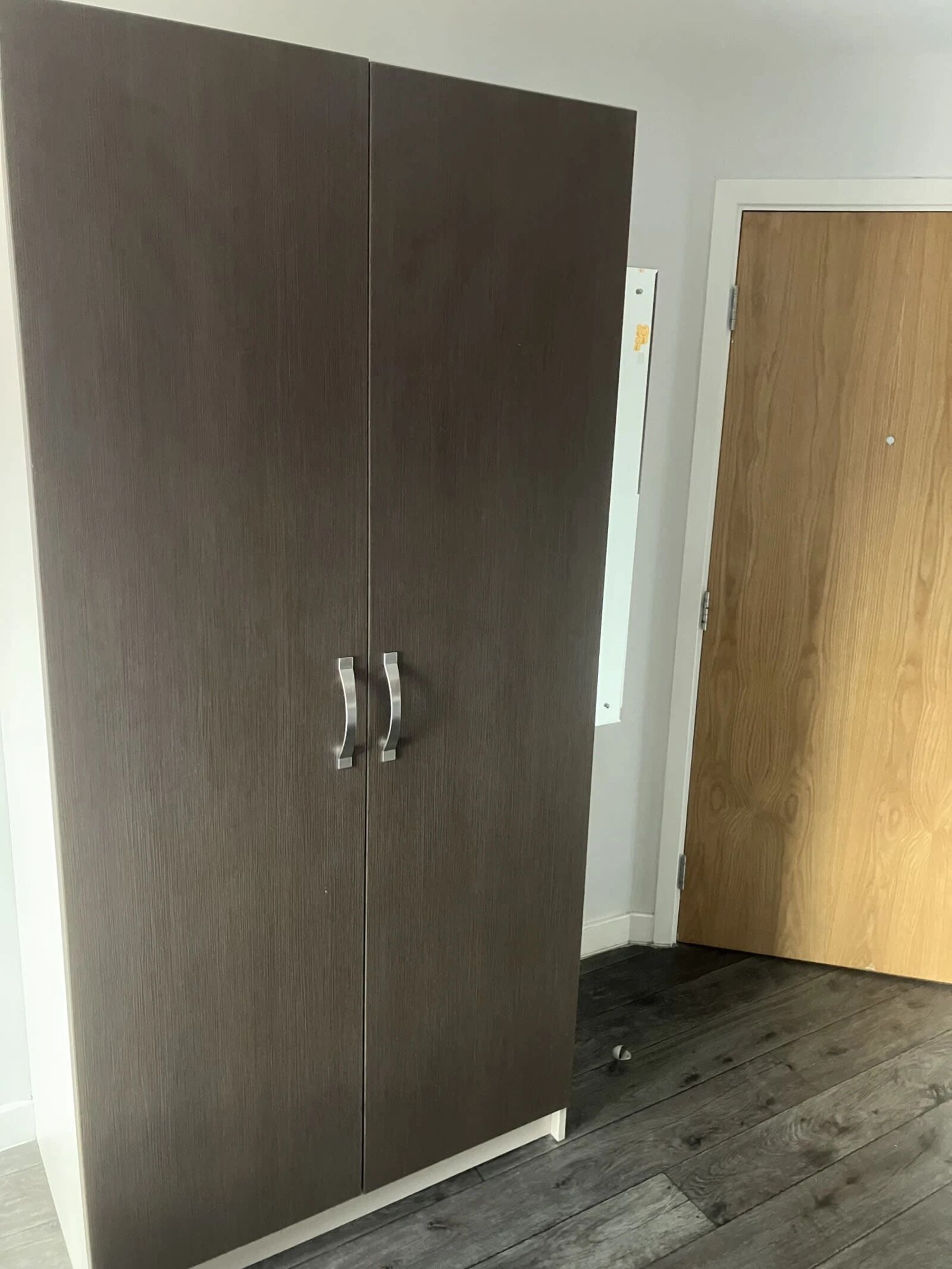 Une armoire en bois sombre, à deux portes et dotée de poignées argentées, est placée à côté d’une porte en bois dans une pièce dont le sol est également en bois.