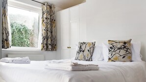 Un lit bien fait, avec du linge de lit blanc et des coussins à motifs floraux, une fenêtre avec des rideaux et un radiateur en dessous.