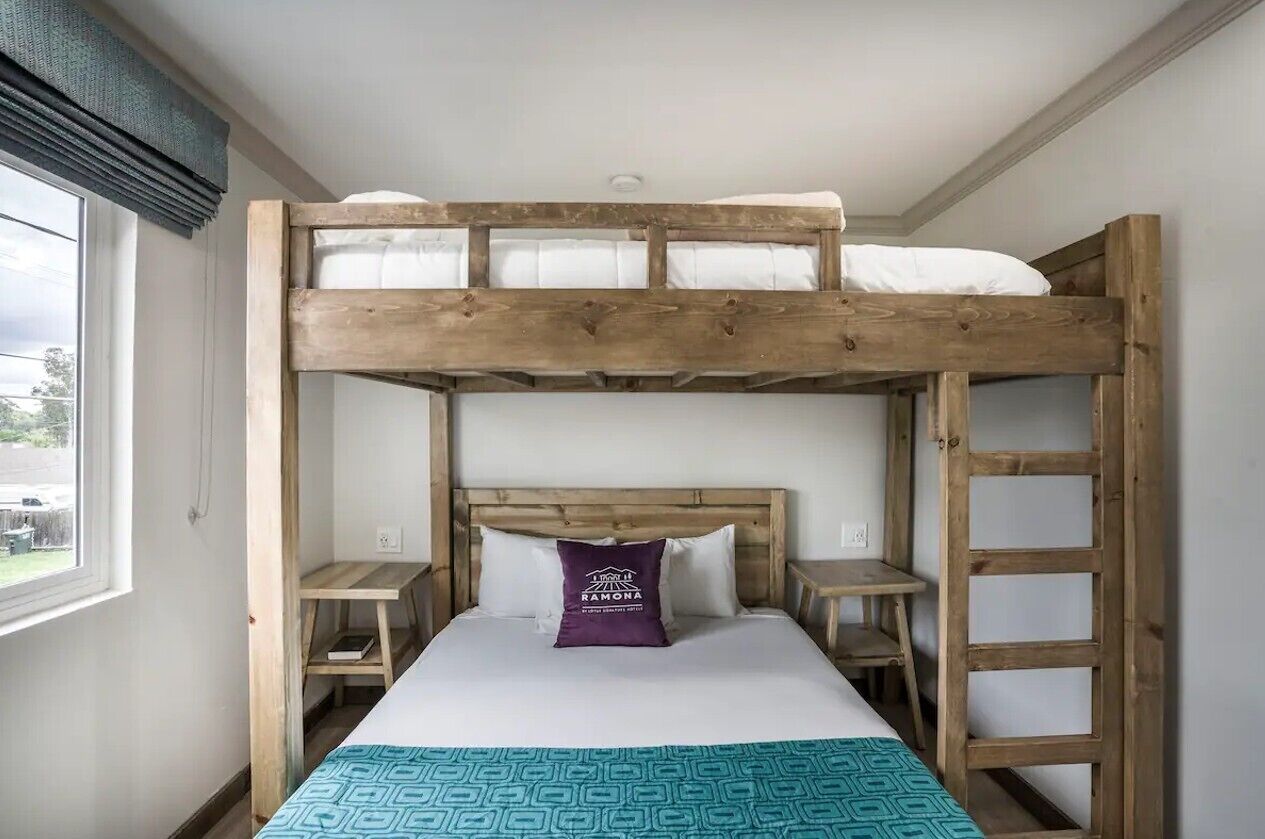 A bunk bed room with a wooden headboard, a purple pillow with text, a small table, and a ladder.