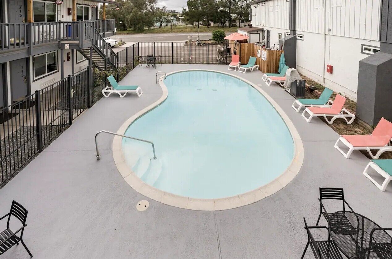 Una piscina con sillas de descanso y una mesita, rodeada por una cerca y un edificio.