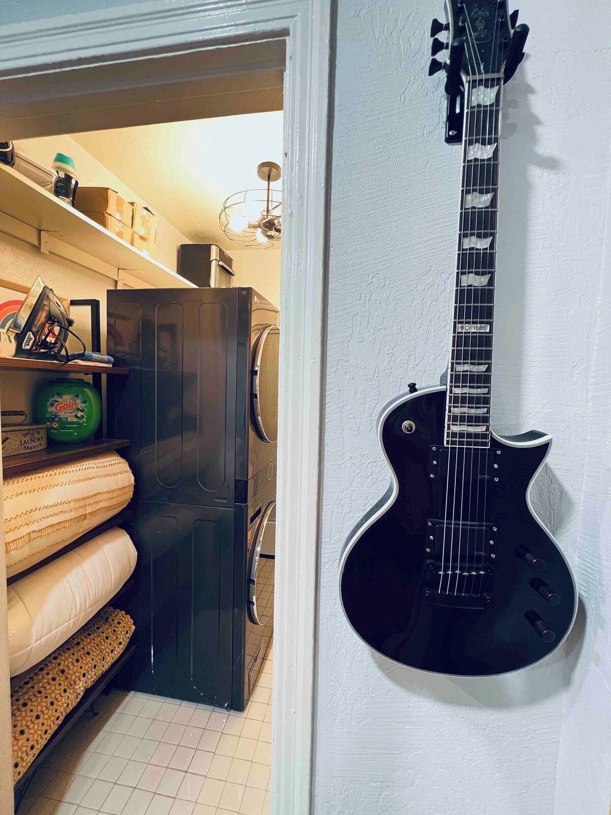 Une pièce dans laquelle on peut voir une guitare noire accrochée au mur, un lave-linge et un sèche-linge dans un coin.