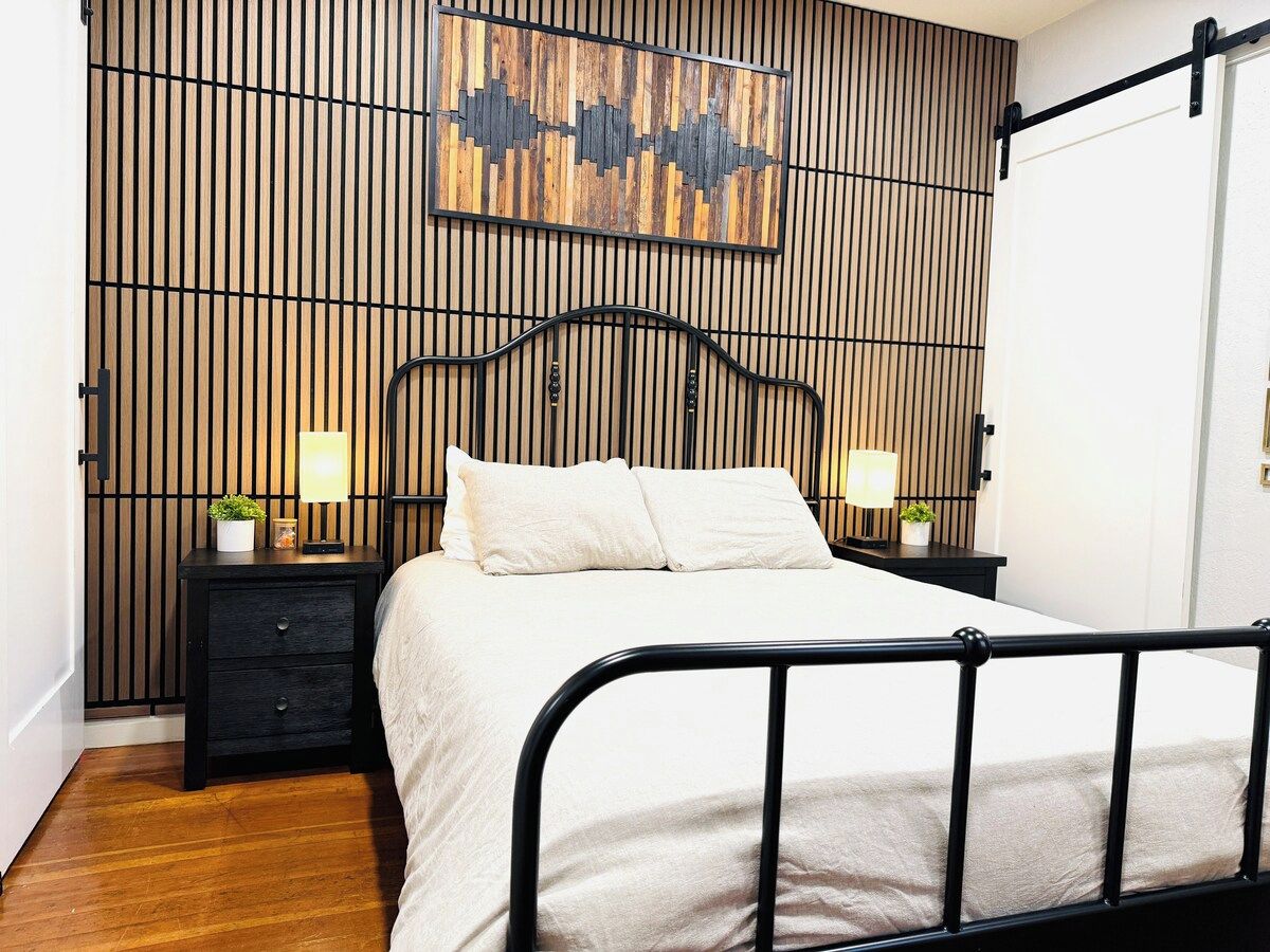 Een slaapkamer met een bed, nachtkastjes, een kunstwerk aan de muur en een zwart metalen hoofdeinde.