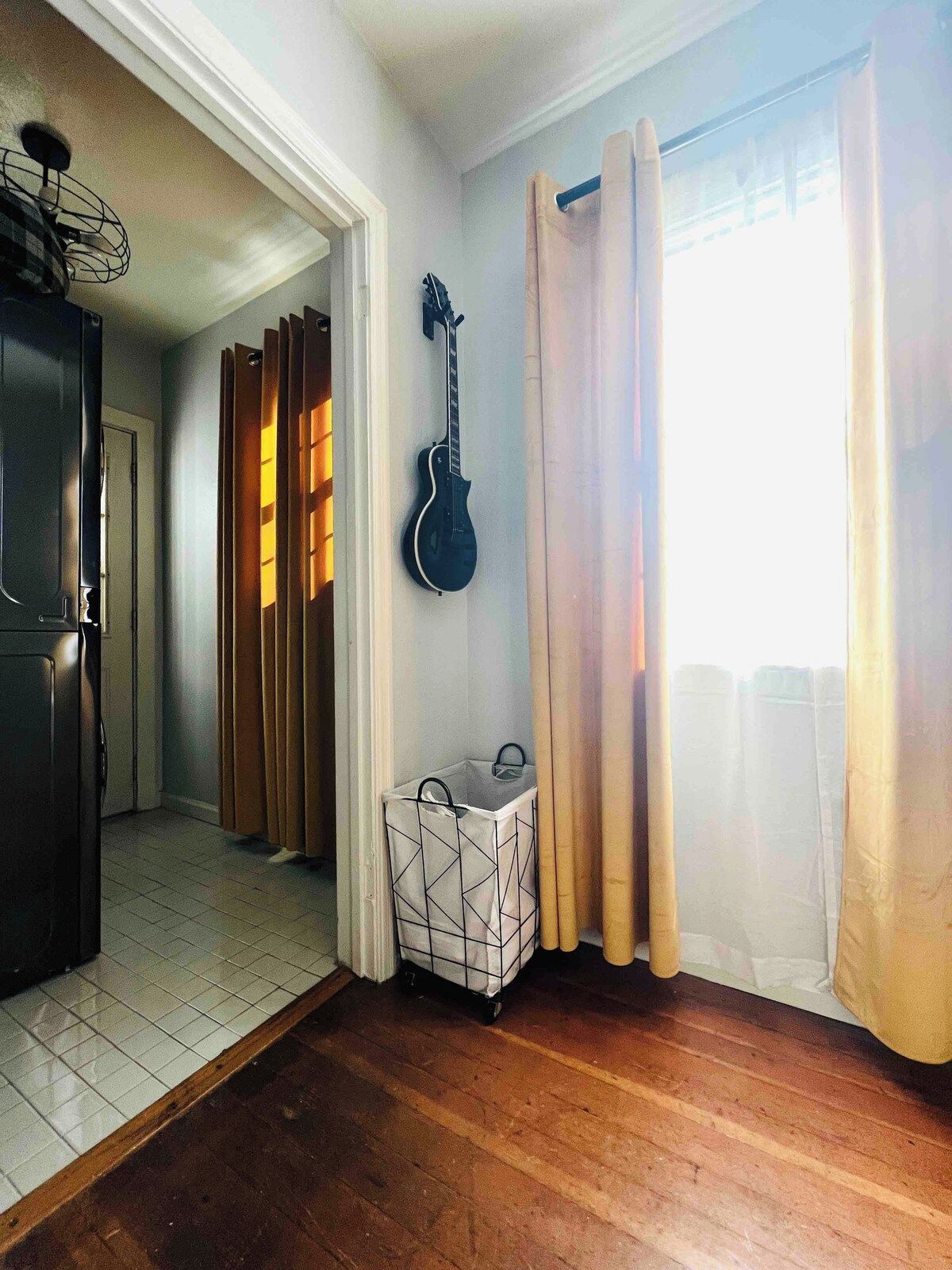 A room with a wooden floor, a guitar hanging on the wall, a storage unit, and a window with curtains.