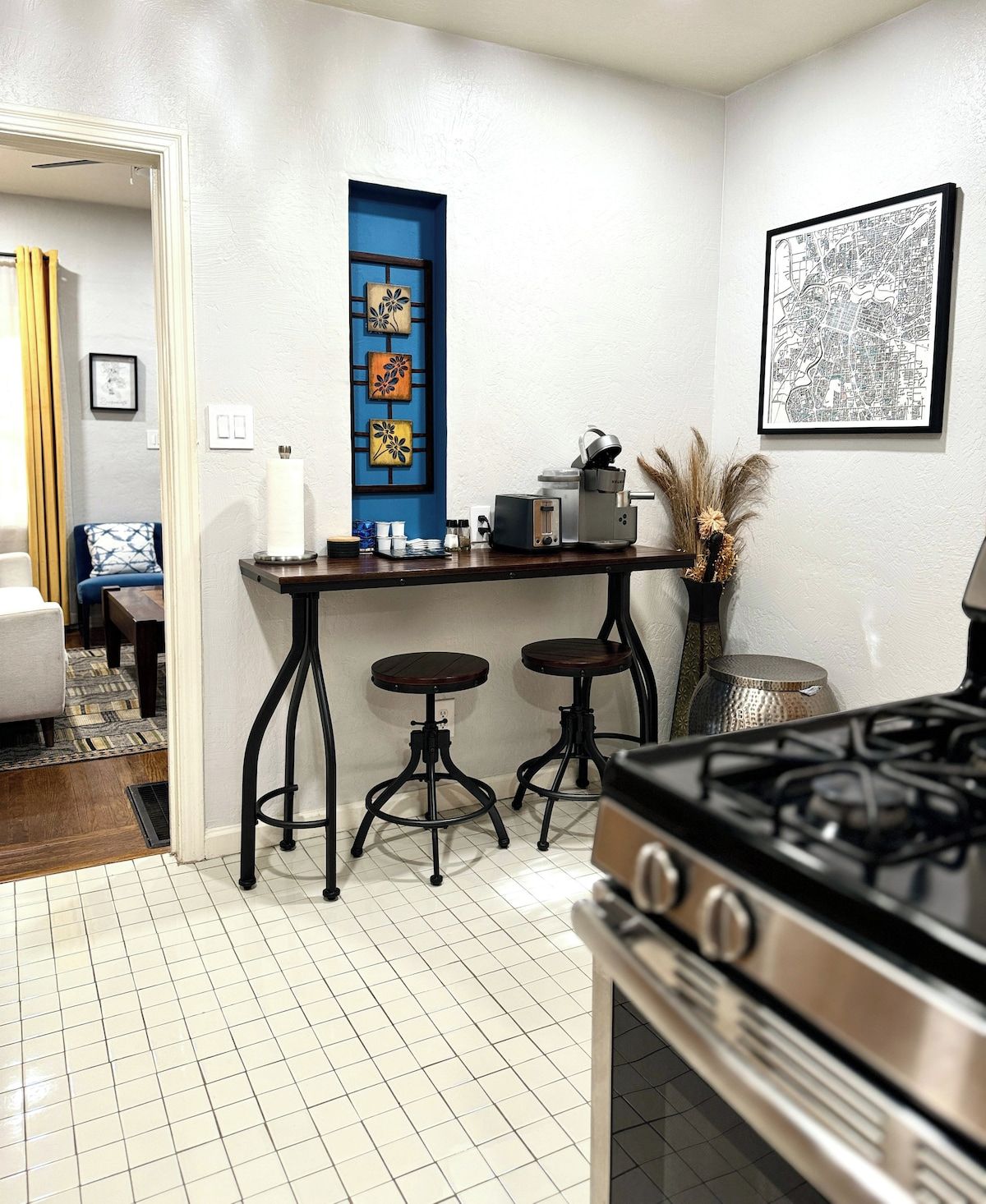 A kitchen with a gas stove, two stools, a coffee machine, and a blue-framed window.