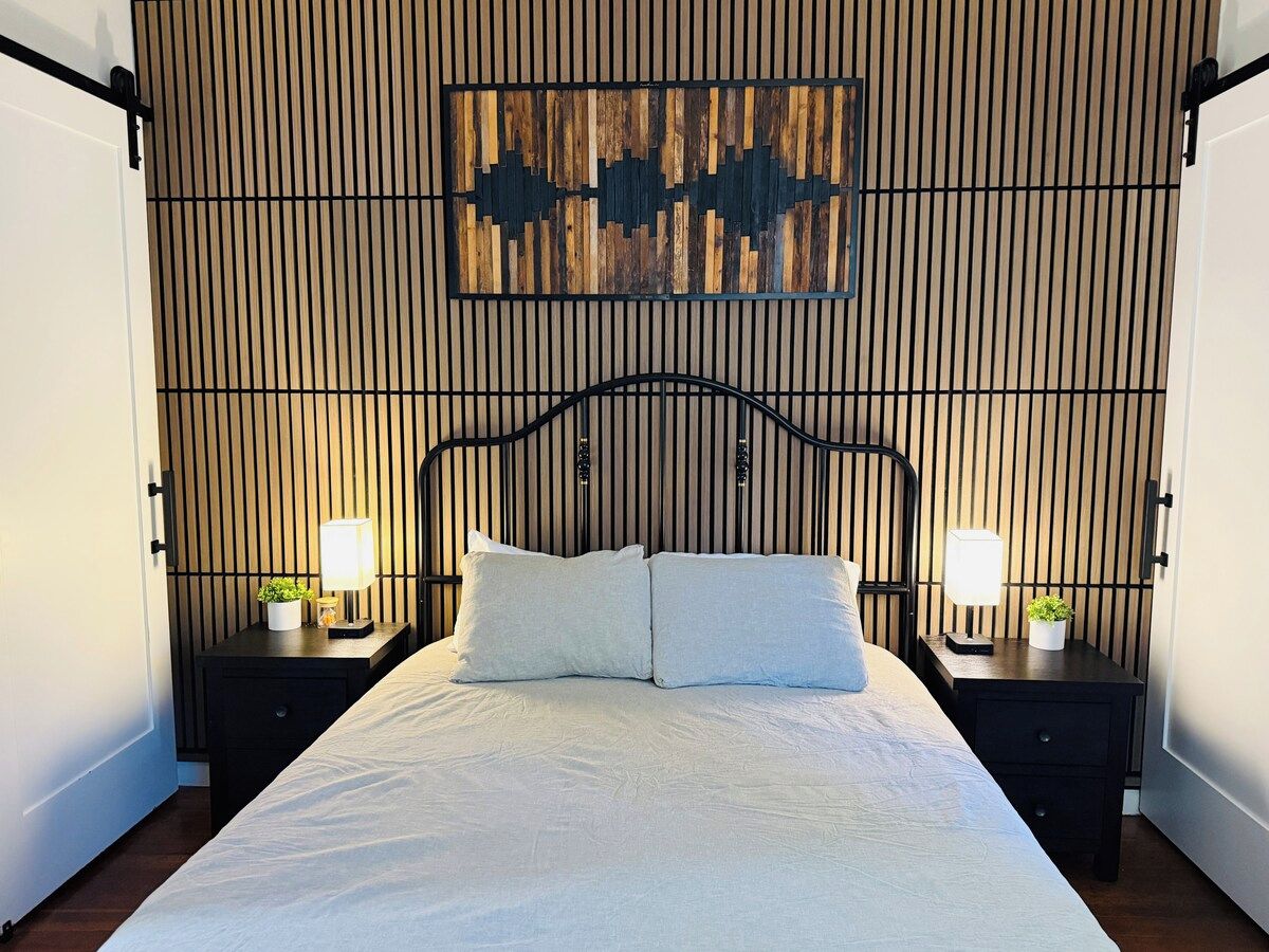 Una cama con cabecera, dos mesitas de noche con lámparas, y una pared con listones de madera verticales y un adorno sobre la cama.